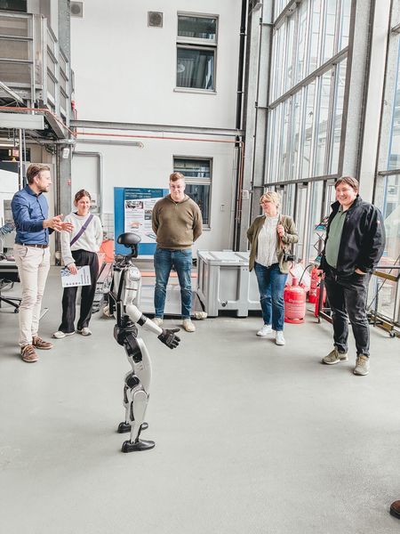 Mehrere Personen stehen in einer hellen Industriehalle um einen humanoiden Roboter, während ein Mann links die Vorführung erklärt.