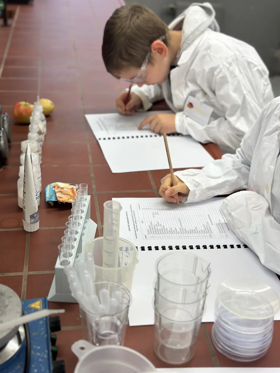KinderUni an der Hochschule Trier in der Lebensmitteltechik - 2 Kinder im Labor notieren etwas in ihr Versuchsskript und haben dabeo Kittel und Schutzbrille an