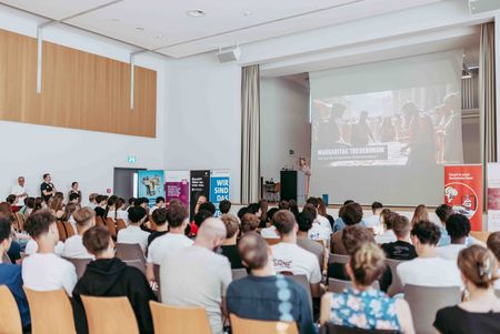 Publikum verfolgt in einem großen Veranstaltungsraum eine Präsentation mit projizierter Folie auf der Leinwand.