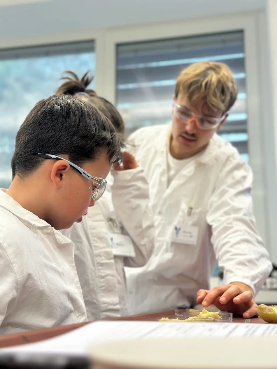 KinderUni an der Hochschule Trier in der Lebensmitteltechik - ein Studierender erklärt zwei Kindern den Versuch, indem er auf eine Petrischale mit geriebenem Apfel zeigt, alle tragen Schutzkleidung