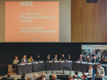 42. Mitgliederversammlung der HRK an der Bauhaus-Universität Weimar; ein großes Podium sitzt vor einer Projektionsfläche mit Veranstaltungsinformationen, während das Publikum im Saal zuhört.