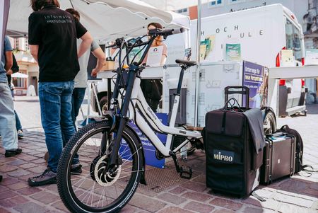 Ein E-Lastenrad steht unter einem Pavillon am Informationsstand der Hochschule Trier. Daneben sind Ausstellungsboxen, Informationsmaterialien und technisches Equipment zu sehen.