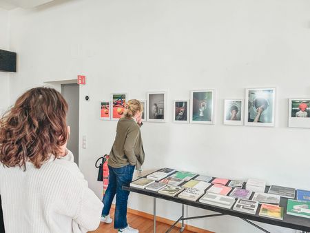 Besucherinnen betrachten gerahmte Fotografien an einer weißen Ausstellungswand; darunter liegen Bücher und Publikationen auf einem langen Tisch.