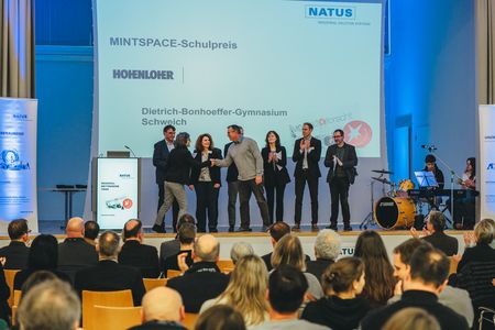 Preisträger*innen stehen auf der Bühne während ein weiterer Betreuer ausgezeichnet wird und die Hadn der Moderatorin schüttelt.