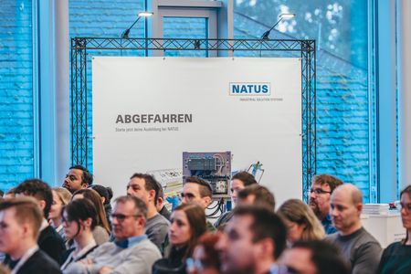 Ein interessiertes Publikum schaut Richtung Bühne während im Hintergrund ein Banner der Natus zu sehen ist.