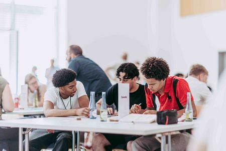 Drei junge Teilnehmer arbeiten konzentriert an einem Tisch und machen sich Notizen. Eine Gruppe junger Menschen steht um einen Tisch und betrachtet Materialien für eine gemeinsame Aufgabe.