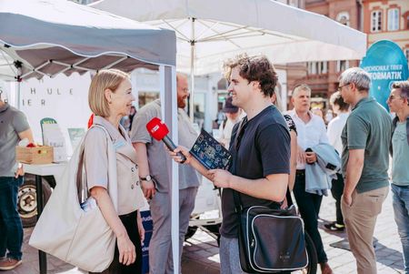 Frau Präsidentin Dorit Schumann wird vor dem Informationsstand der Hochschule Trier von einem Mann mit Mikrofon interviewt. Im Hintergrund sprechen weitere Besucherinnen und Besucher miteinander.