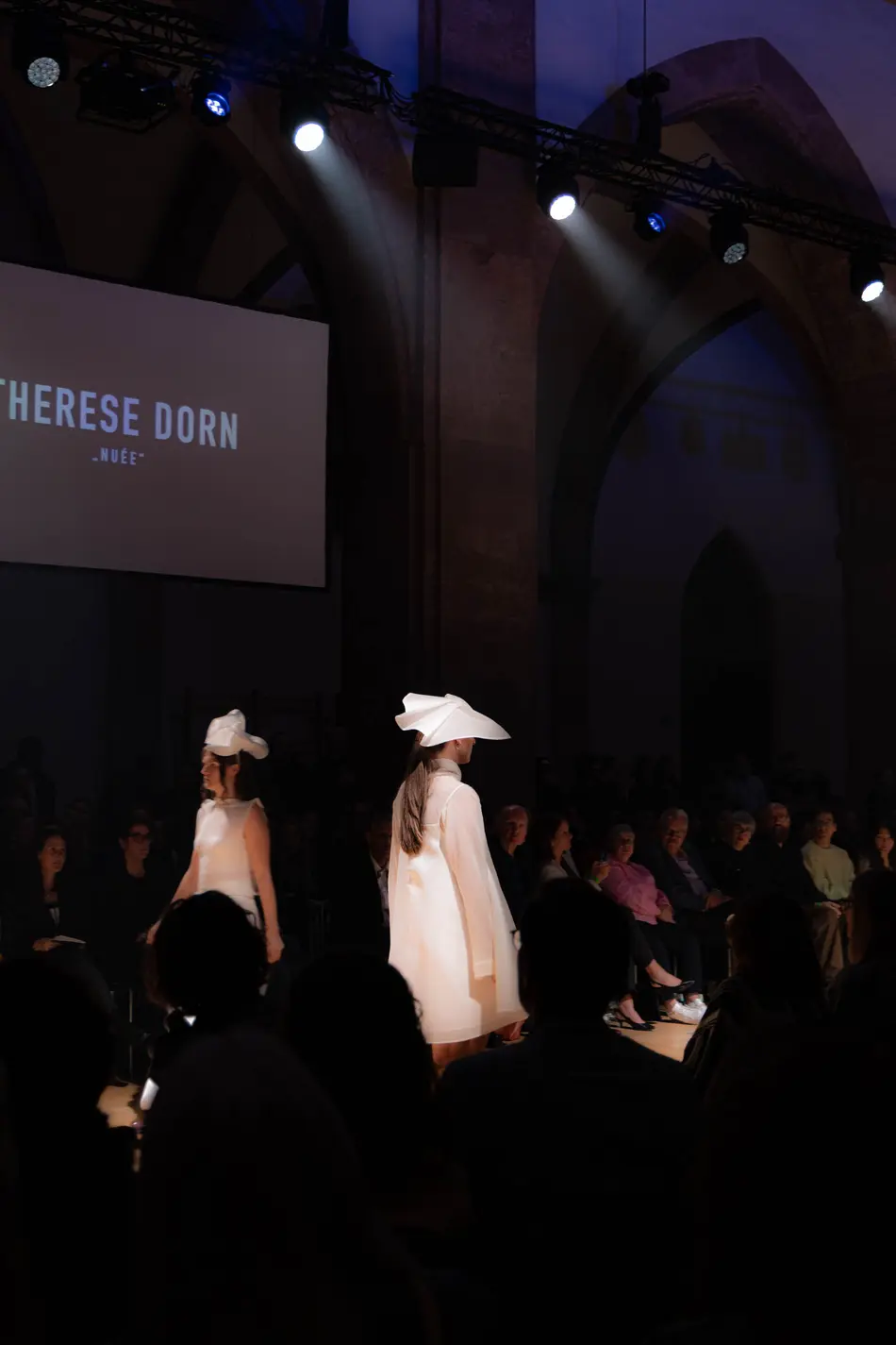 Zwei Models zeigen weiße Outfits mit auffälligen, skulpturalen Kopfbedeckungen.
