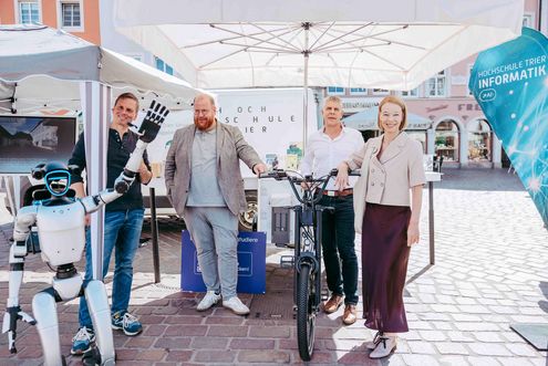 Vier Personen posieren unter einem Pavillon am Informationsstand der Hochschule Trier neben einem E-Lastenrad. Links im Bild hebt ein humanoider Roboter die Hand.