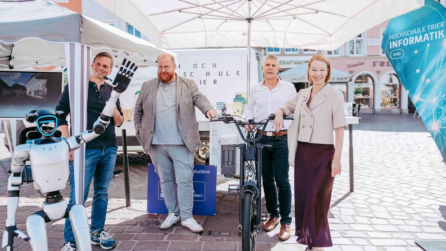 Vier Personen posieren unter einem Pavillon am Informationsstand der Hochschule Trier neben einem E-Lastenrad. Links im Bild hebt ein humanoider Roboter die Hand.