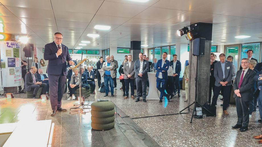 Empfangssituation mit Rede: Ein Mann spricht mit Mikrofon vor einem Publikum in einem hellen Foyer, umgeben von Stehtischen, Veranstaltungstechnik und Zuhörenden.