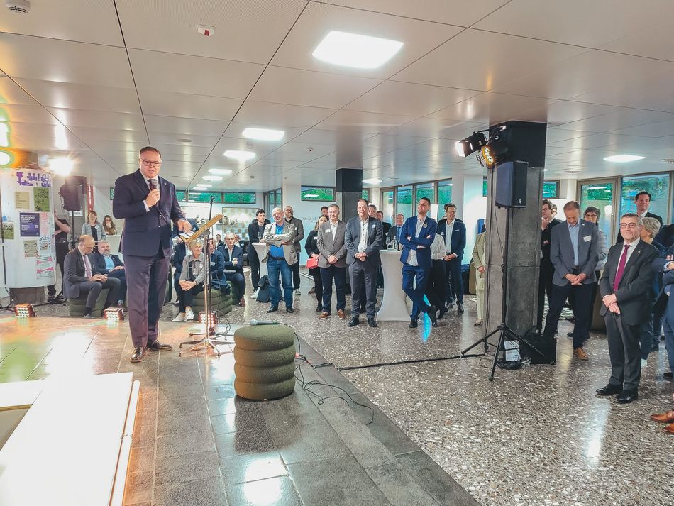 Empfangssituation mit Rede: Ein Mann spricht mit Mikrofon vor einem Publikum in einem hellen Foyer, umgeben von Stehtischen, Veranstaltungstechnik und Zuhörenden.