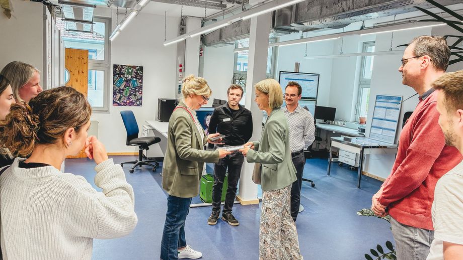 Eine Gruppe von Besucherinnen und Besuchern steht in einem Laborraum, während zwei Frauen in der Mitte Unterlagen oder Exponate austauschen.