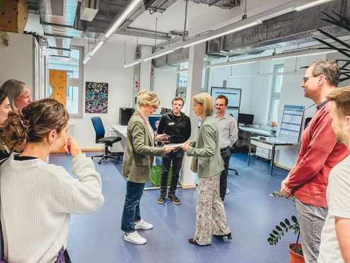 Eine Gruppe von Besucherinnen und Besuchern steht in einem Laborraum, während zwei Frauen in der Mitte Unterlagen oder Exponate austauschen.