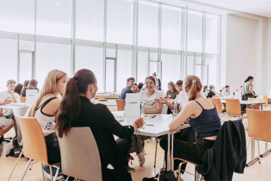 Studierende sitzen in Gruppen an Tischen in einem hellen Seminarraum und arbeiten gemeinsam an einer Aufgabe.