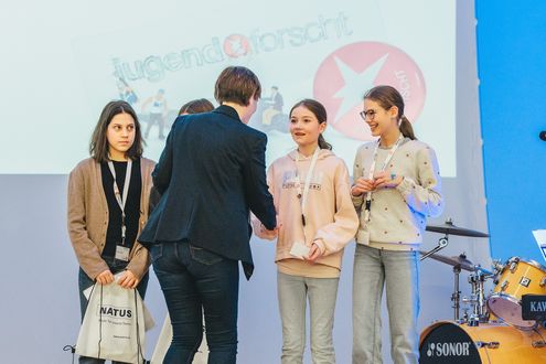 Mehrere Kinder und Jugendliche stehen auf einer Bühne, während einer Person aus dem Organisationsteam eine Urkunde überreicht wird; im Hintergrund ist das ‚Jugend forscht‘-Logo zu sehen.