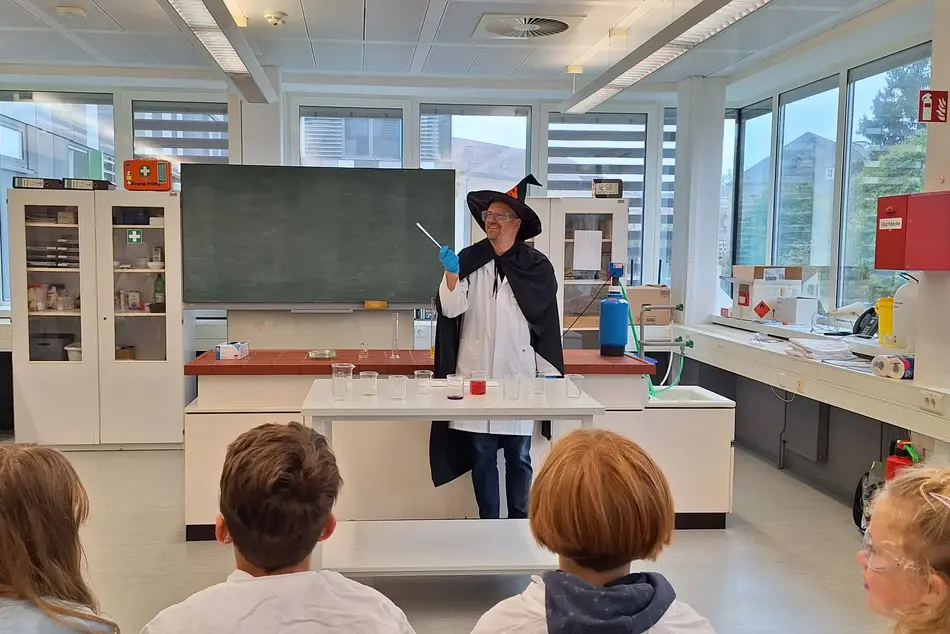 KinderUni an der Hochschule Trier in der Lebensmitteltechik - Prof. Dr. Heiko Oertling als Zauberer verkleidet führt ein Experiment vor, vor ihm stehen Bechergläser teilweise gefüllt mir farbigen oder farblosen Lösungen
