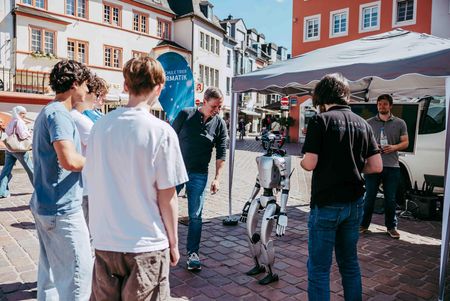 Mehrere Besucherinnen und Besucher stehen vor dem Informationsstand der Hochschule Trier auf einem Stadtplatz und interagieren mit einem humanoiden Roboter.