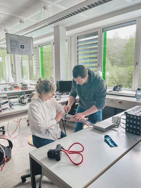 Zwei Personen stehen in einem Techniklabor an einem Tisch und begutachten gemeinsam eine elektronische Komponente; im Vordergrund liegen Kamera, Kabel und technische Geräte.