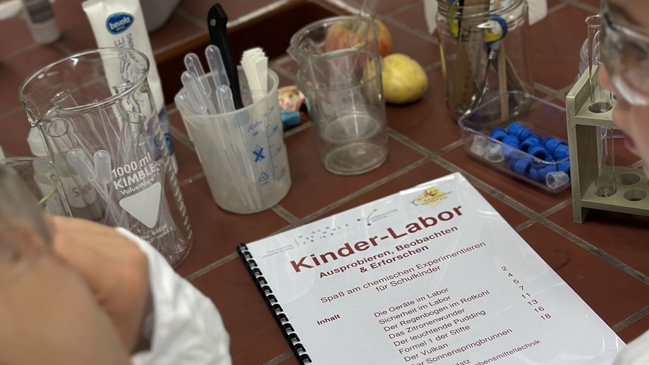 KinderUni an der Hochschule Trier in der Lebensmitteltechnik - das Versuchsskript des Kinder-Labors liegt auf der Laborbank zwischen Kindern in Kittel und Schutzbrille