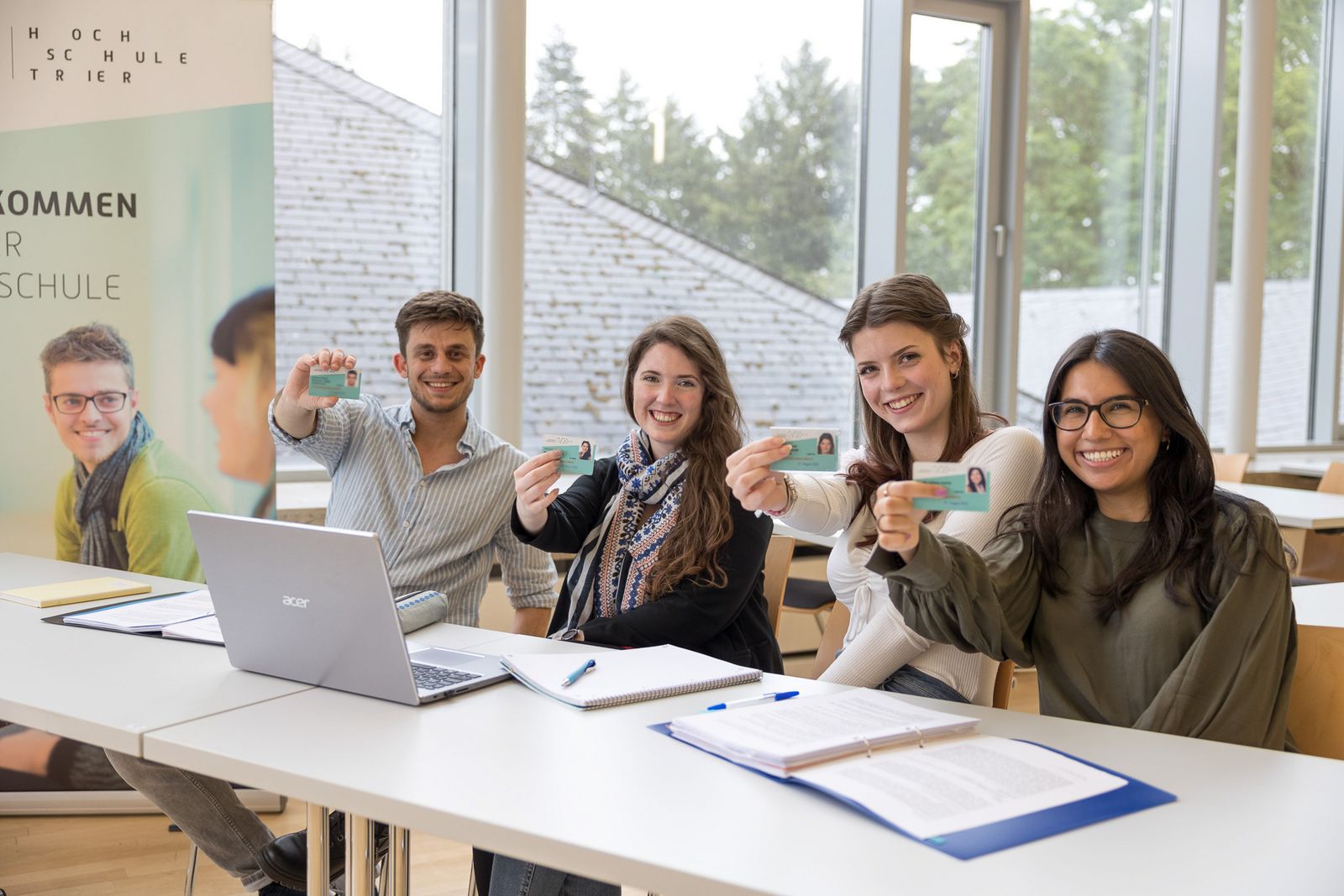 Vier Studierende sitzen lächelnd an einem Tisch in einem hellen Seminarraum und halten ihre Studierendenausweise in die Kamera; auf dem Tisch liegen ein Laptop, Notizblock und Unterlagen, daneben steht ein Banner der Hochschule Trier.