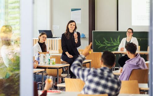Menschen sitzen in einem hellen Klassenraum an Tischen, während eine Frau vorne spricht und ein Glas hochhält; im Vordergrund meldet sich eine Person.