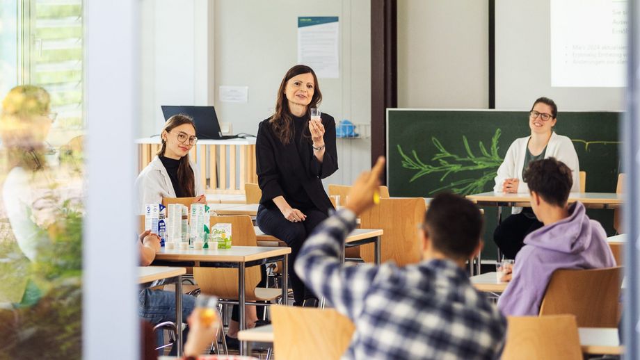 Menschen sitzen in einem hellen Klassenraum an Tischen, während eine Frau vorne spricht und ein Glas hochhält; im Vordergrund meldet sich eine Person.