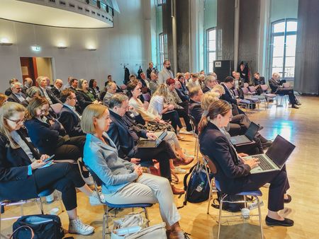 Teilnehmende einer HRK-Sitzung sitzen in einem Veranstaltungssaal; einige arbeiten an Laptops oder hören einem stehenden Redner im Publikum zu.