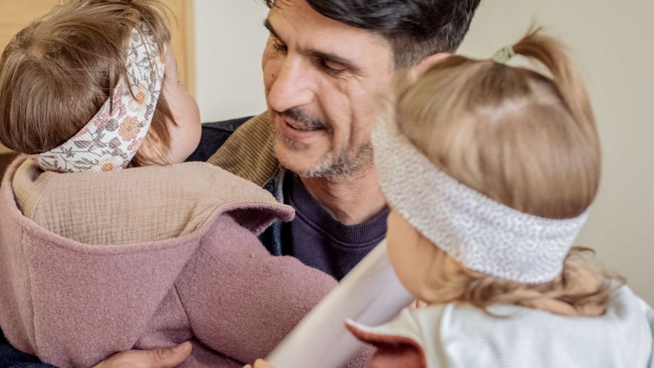 Ein Vater hält seine zwei kleinen Kinder auf dem Arm und lächelt ihnen zu. Die Kinder tragen Stirnbänder und sind ihm zugewandt.
