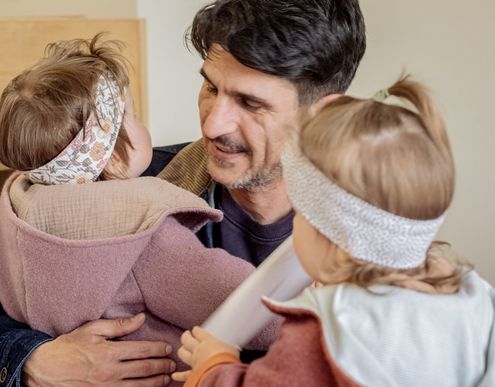 Ein Vater hält seine zwei kleinen Kinder auf dem Arm und lächelt ihnen zu. Die Kinder tragen Stirnbänder und sind ihm zugewandt.